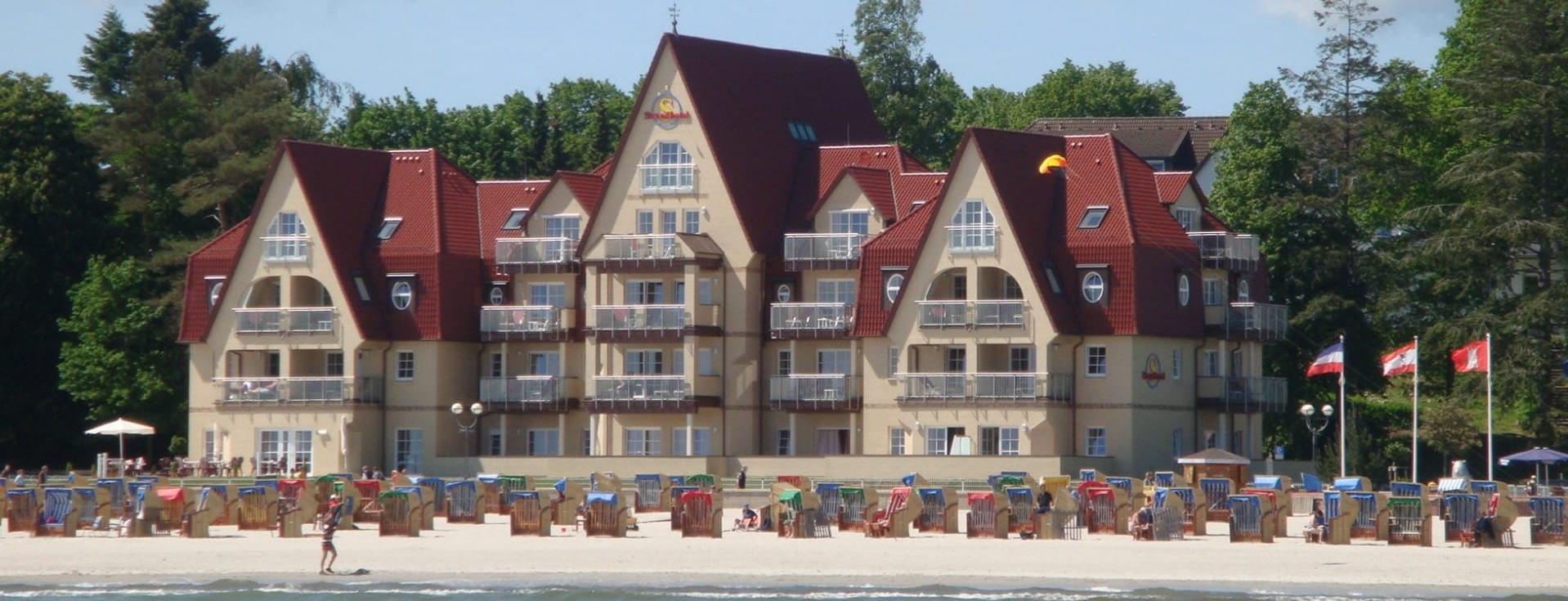 Ein Hotelgebäude im Stil eines Ferienhauses mit rotem Dach, direkt am Strand gelegen. Im Vordergrund ist der Sandstrand sichtbar, der von möglichst vielen Sonnenliegen umgeben ist. Die einladende Szenerie deutet auf einen Freizeitort hin.