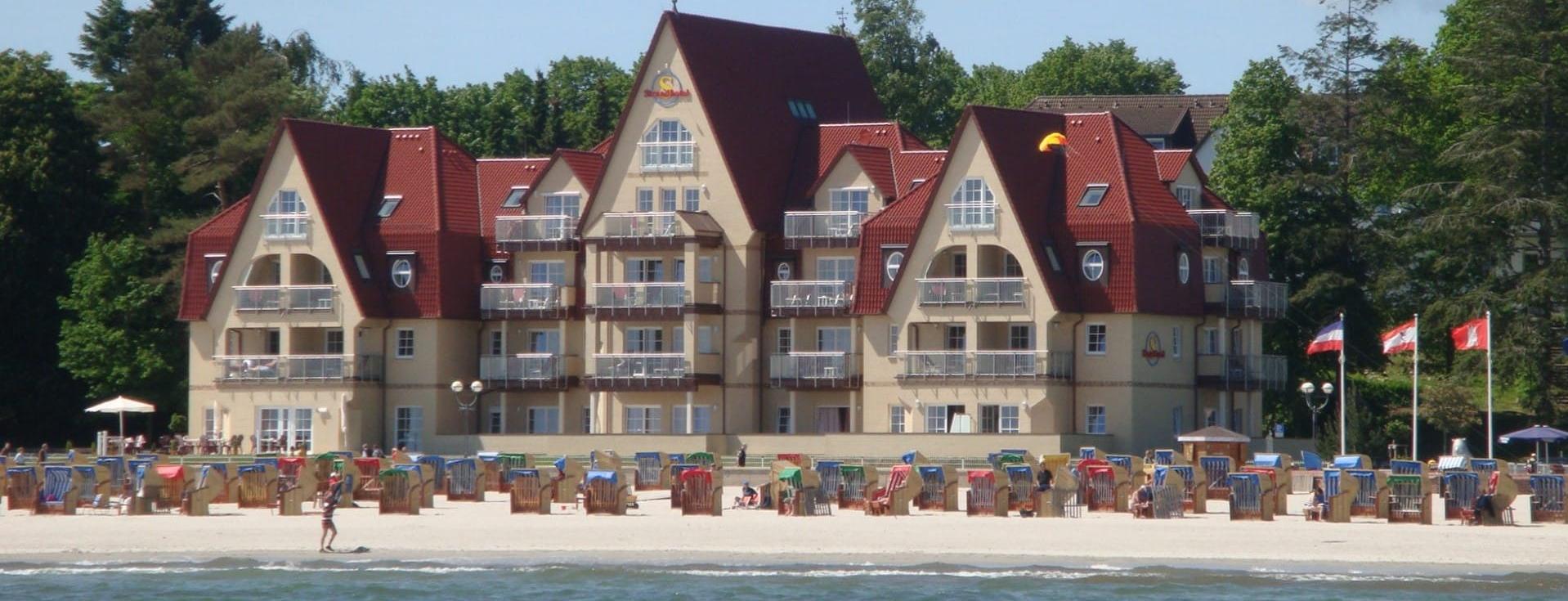 Ein Hotelgebäude im Stil eines Ferienhauses mit rotem Dach, direkt am Strand gelegen. Im Vordergrund ist der Sandstrand sichtbar, der von möglichst vielen Sonnenliegen umgeben ist. Die einladende Szenerie deutet auf einen Freizeitort hin.